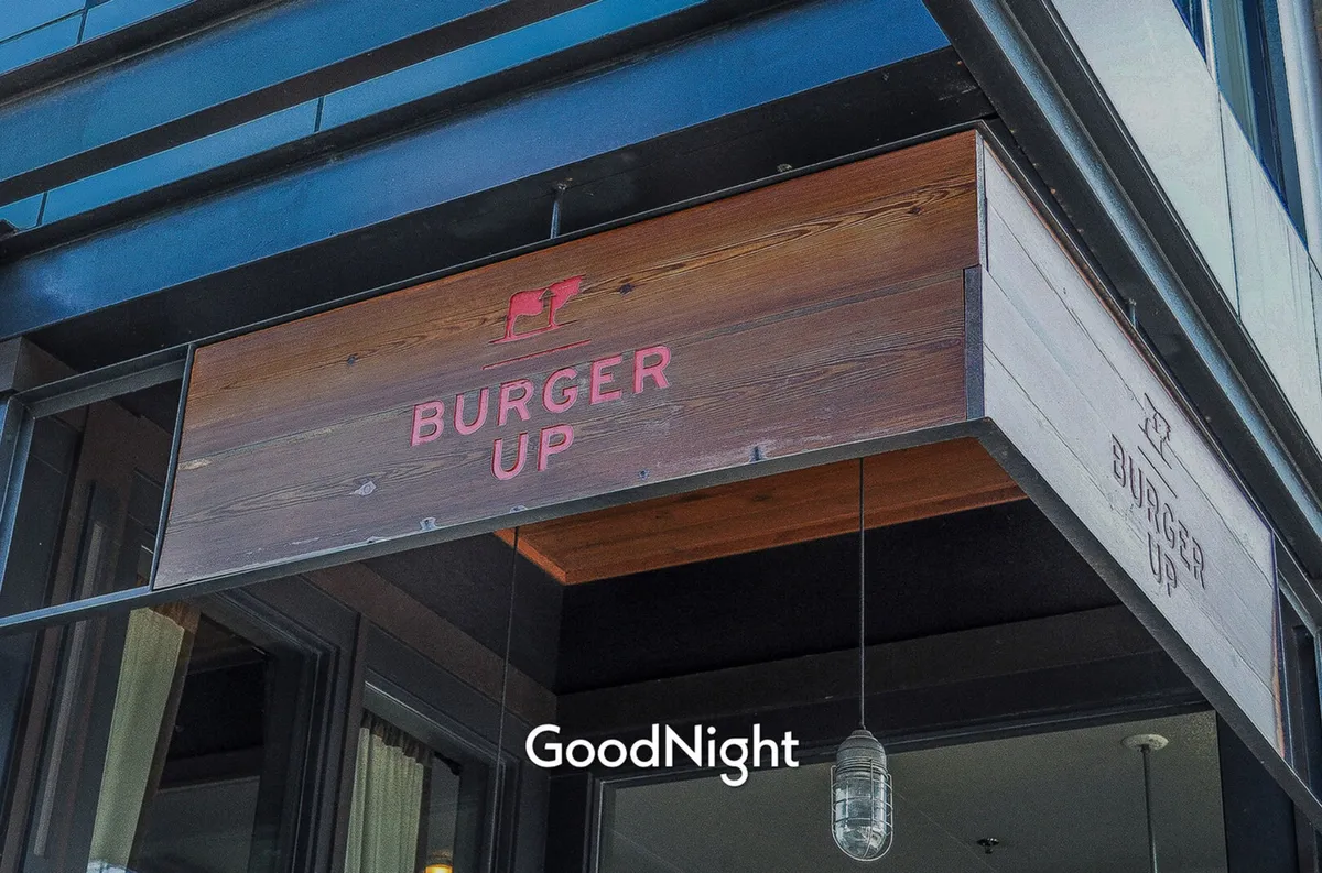 The distinctive Burger Up sign hangs above a warmly lit entrance, shaded by a hanging lamp. The scene captures the inviting ambiance of one of the district's beloved eateries, setting the tone for casual gatherings and gourmet burgers.