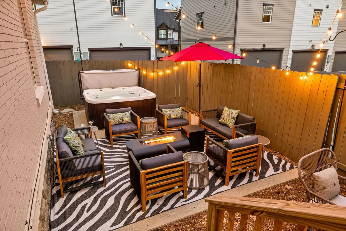 An inviting outdoor seating area featuring a hot tub surrounded by string lights, a comfortable seating arrangement, a zebra-patterned rug, and a red umbrella. The scene is designed for relaxation and evening gatherings, exuding a cozy, intimate atmosphere.
