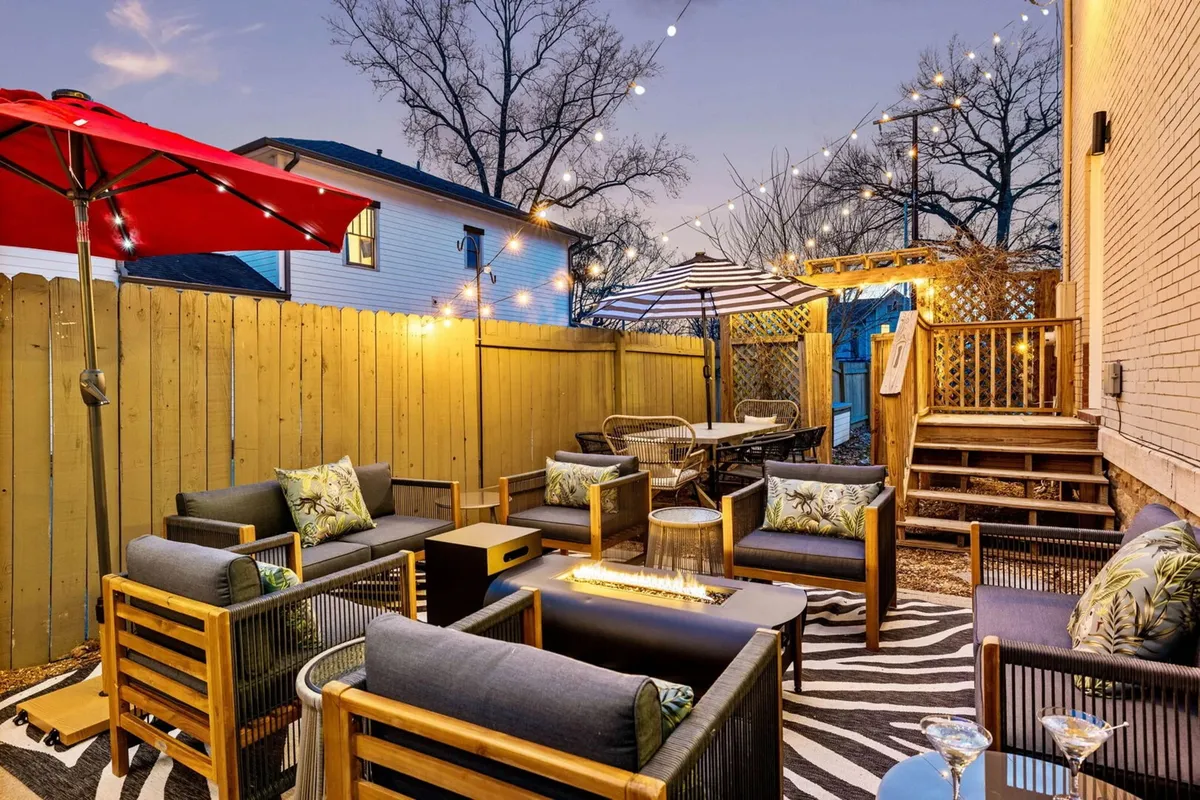 A cozy outdoor seating area featuring a table, comfortable chairs, string lights overhead, an umbrella for shade, and a wooden privacy fence enclosing the space. The ambiance is warm and inviting, ideal for relaxed gatherings or intimate conversations as the sun sets.