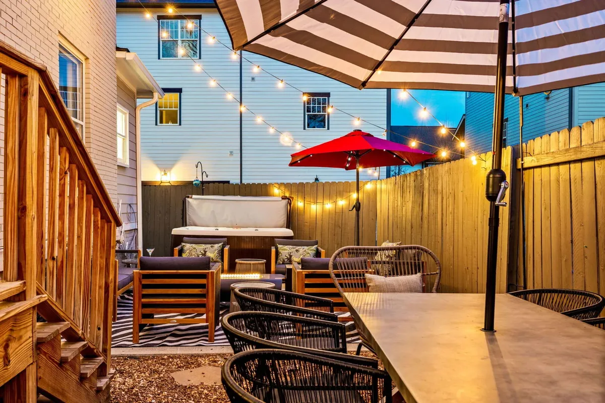 A festive outdoor patio space with striped umbrellas, string lights, a hot tub, and ample seating. The setup is ideal for socializing, dining al fresco, or enjoying a relaxing soak under the stars.