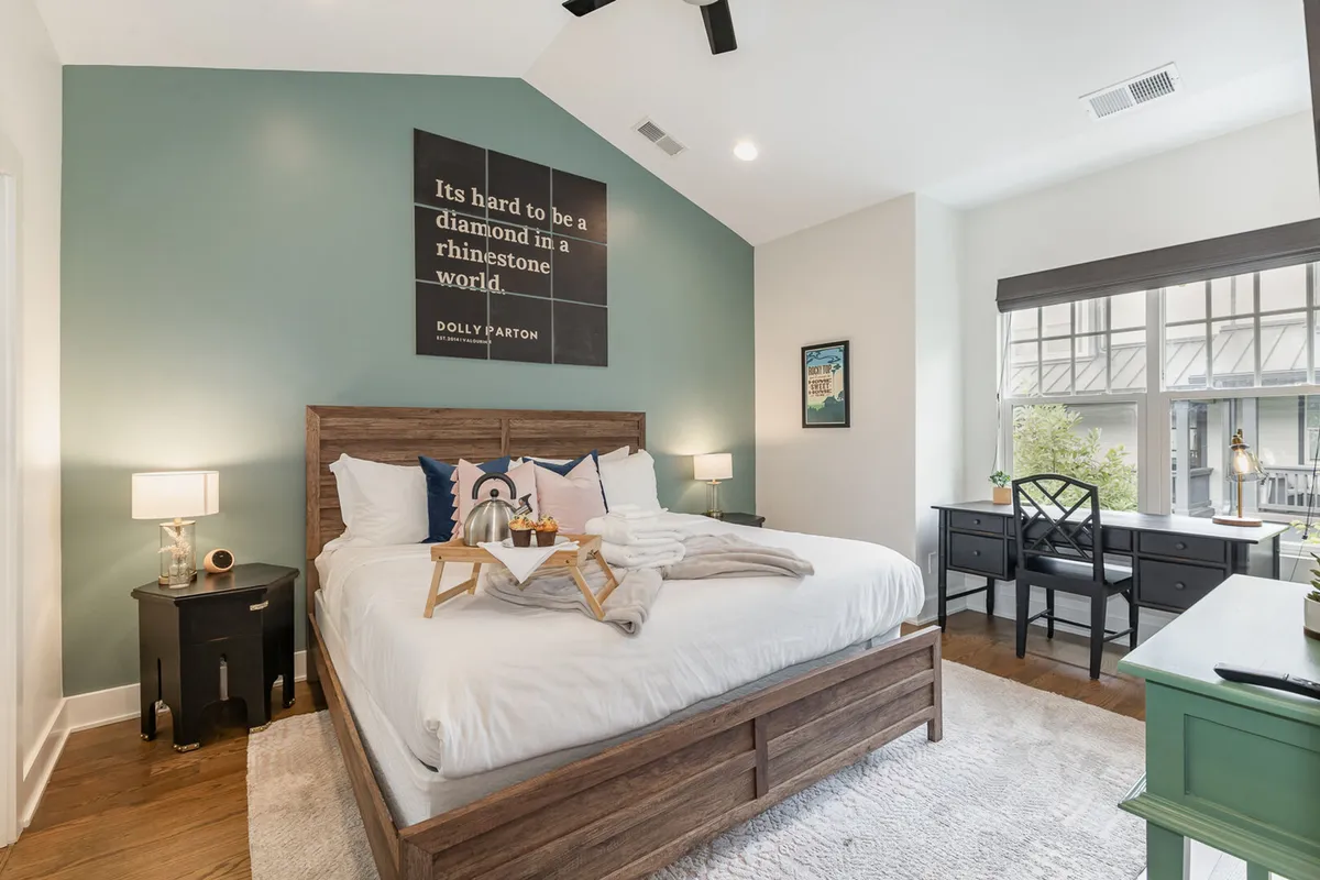 A serene bedroom featuring a plush bed with crisp white linens and decorative pillows, Dolly Parton quote art above the headboard, a cozy desk area by the window, and abundant natural light creating a peaceful ambiance.