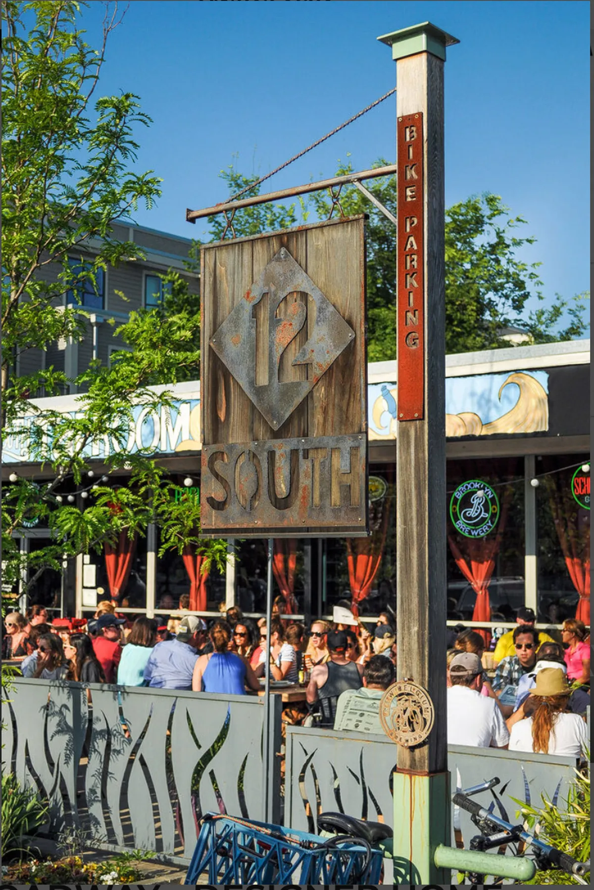This vibrant street scene features the iconic 12 South sign, lively outdoor seating with diners, bike parking signage, and decorative railings. Locals and visitors are enjoying the sunshine, reflecting the neighborhood's walkable, energetic vibe.