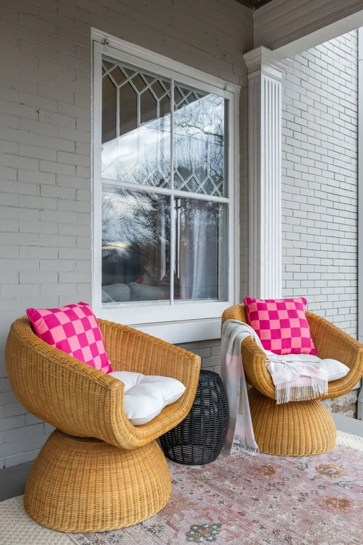 A relaxed outdoor nook with wicker chairs adorned with colorful pillows, a decorative rug, a black side table, and a window set into the house. The space basks in natural light and is designed for coffee sipping, reading, or quiet, intimate moments outdoors.