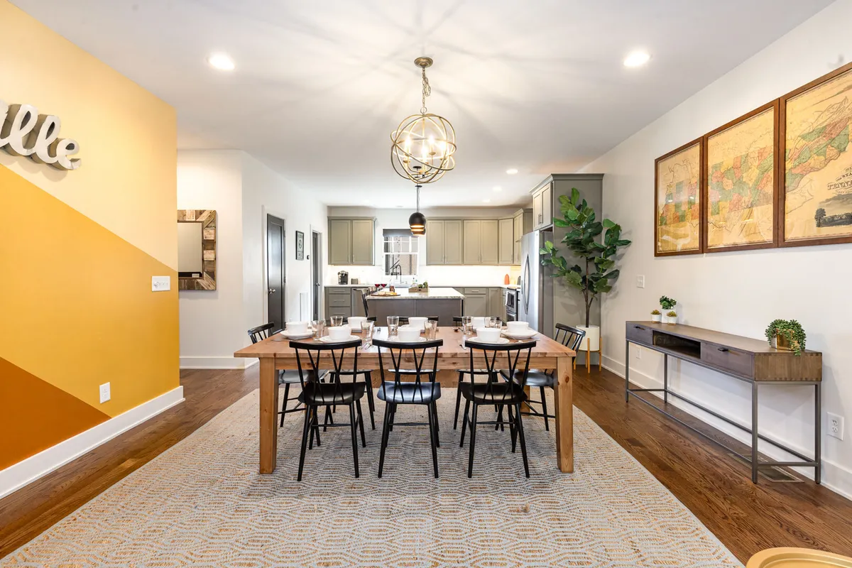 A warm, inviting dining area set with a large wooden table, contemporary chandelier, and adjacent kitchen space. Wall art and a potted plant add a homey touch, setting the stage for communal meals and celebrations.