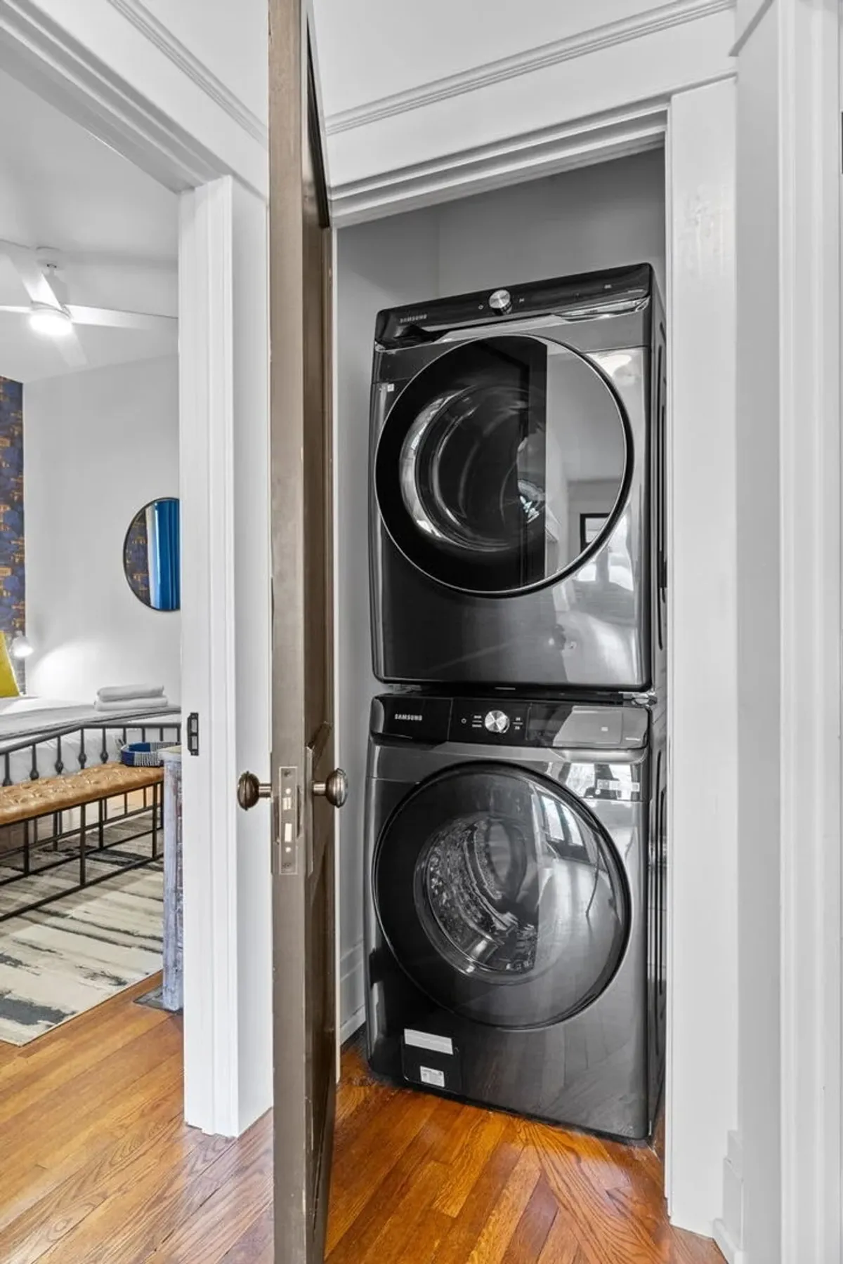 This utility space features a stacked washer and dryer set against a clean, modern backdrop. The wooden flooring and adjacent doorway make it an accessible and practical amenity within the home.